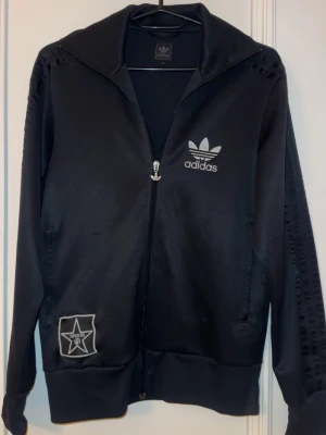 Svart zip hoodie från Adidas - Snygg svart hoodie från Adidas med dragkedja framtill och klassisk Adidas-logga på bröstet. Tröjan har långa ärmar, ribbade muddar och en stjärnpatch på ena sidan. Hoodien ger en sportig/chill vibe. Pris går att diskutera. 💞 