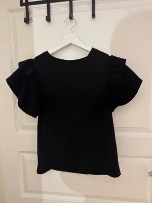 Svart topp med volangärmar - Snygg svart topp med stora volangdetaljer på ärmarna. Rak passform och rund halsringning. Perfekt för dig som vill ha en enkel men ändå trendig look. Passar till både jeans och kjol.