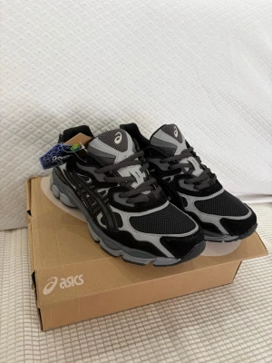 Svarta och grå Asics sneakers - Snygga Asics sneakers i svart och grått med meshpaneler och detaljer i syntet. Skorna har en sportig look med klassisk Asics-logga på sidan och tungan. Sulan är robust och ger bra stöd, perfekt för dig som gillar streetwear och sportig stil.