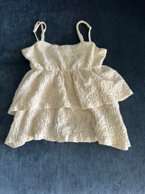 Beige volangtop med smala axelband - Supersöt beige linnetopp med volanger och smala axelband. Toppen har en skrynklig struktur och är ärmlös, perfekt för varma dagar. Lätt och luftig modell som ger en romantisk känsla.