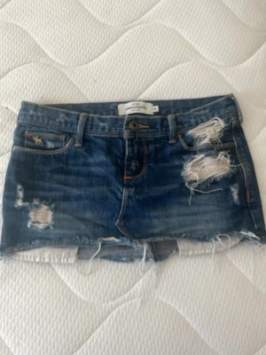 Blå sliten jeanskjol Abercrombie - Säljer ett par blå jeanskjol från Abercrombie & Fitch med slitna detaljer och råa kanter. Klassisk femficksmodell med låg midja och broderad logga på fickan. Perfekta för en avslappnad och trendig sommarstil.  Midjemått:35.5cm längd:20.5 cm