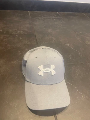 Ljusblå keps från Under Armour - Snygg ljusblå keps från Under Armour med böjd skärm och klassisk passform. Kepsen har ett broderat vitt logotypmärke framtill och ventilationshål på ovansidan. Tillverkad i ett tåligt tyg som passar perfekt till en sportig stil.