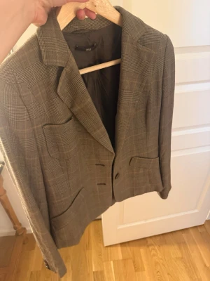 Rutig brun kavaj med mockadetaljer - Snygg brunrutig kavaj med klassiskt slag och två framfickor. Kavajen har mockadetaljer på armbågarna och stängs med två knappar framtill.