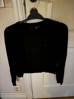 Svart kort kofta - En svart, kort kofta från 157 med rund halsringning och knappar framtill. Koftan har långa ärmar och ribbade muddar vid ärmslut och nederkant. Enkel och stilren design som passar till många olika outfits.