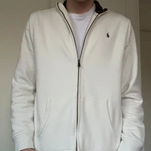 Vit hoodie från Polo Ralph Lauren - Säljer en stilren vit hoodie från Polo Ralph Lauren med dragkedja och broderad logga i blått. Den har en rutig krage i rött och grönt som ger en snygg kontrast. Perfekt för en avslappnad look. Använd bara ett fåtal gånger. Pris kan diskuteras. Passar xs-S