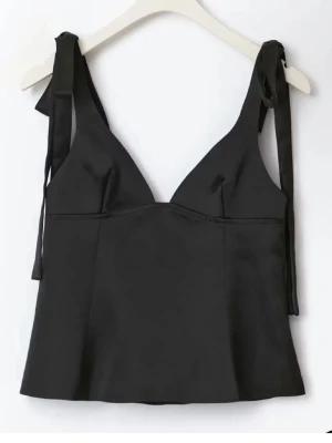 Svart satinlinnetopp med knytband - Stilren svart linnetopp i satin med smickrande v-ringning och breda axelband som knyts på axlarna. Toppen har en lätt glansig finish och är perfekt för att skapa en chic look. Passar till både jeans och kjol.