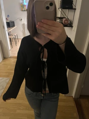 Svart knytkofta med vida ärmar - Trendig svart kofta med knytning framtill och vida ärmar. Koftan har en croppad passform och är tillverkad i ett mjukt, tunt material. Perfekt att styla över ett linne eller topp för en avslappnad look.