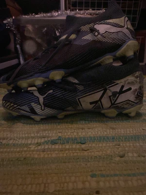 Puma Future blåa fotbollsskor - Säljer ett par blåa Puma Future fotbollsskor med vita och svarta detaljer samt logga på sidan. Skorna har en strumpliknande passform och fasta dobbar i ljusgul färg, perfekta för spel på gräsplan. Snygg design med grafiska mönster och slitstarkt syntetmaterial.