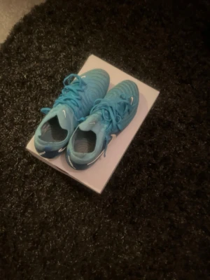 Blå Nike fotbollsskor med snörning - Säljer ett par blå Nike fotbollsskor med klassisk snörning och vit swoosh på sidan. Skorna har en modern, sportig design och är tillverkade i syntetmaterial som ger bra grepp och komfort på planen. Perfekta för dig som vill sticka ut på matchen!