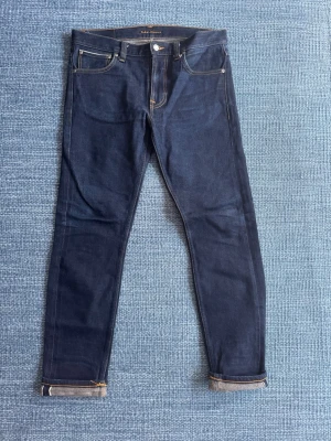 Nudie jeans dry true selvedge denim  - Ett par i princip nya Lean Dean selvedge-jeans från Nudie Jeans i mycket fint skick.  Perfekta för dig som vill skapa ett par jeans med egen karaktär – ju mer de används desto snyggare blir de.  Nypris ligger kring ca 2000 kr.  Mått (Ca) : Total längd: 99 cm Innerbens längd: 74 cm Midja: 43 cm Benöppning: 16 cm  Hör gärna av dig vid frågor eller om du vill ha fler bilder! 