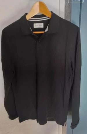 Svart långärmad pikétröja Pier One - Klassisk svart långärmad pikétröja från Pier One med krage och knäppning framtill. Tröjan har en stilren look och är perfekt för en avslappnad men ändå uppklädd stil. Tillverkad i mjukt material för extra komfort.