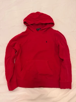 Röd hoodie från Polo Ralph Lauren - Snygg röd hoodie från Polo Ralph Lauren med klassisk huva och känguruficka framtill. Liten mörkblå logga broderad på bröstet. Tillverkad i mjukt bomullsmaterial och har ribbade muddar vid ärmslut och nederkant. Perfekt för en avslappnad och stilren look.
