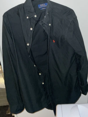 Svart skjorta från Polo Ralph Lauren - Klassisk svart skjorta från Polo Ralph Lauren med vit knappstängning och röd broderad logga på bröstet. Den är tillverkad i bomull och har en normal passform. Nypris 1800 kr. Pris går att diskuteras!