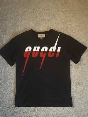 Svart Gucci t-shirt med röd/vit print - Snygg svart t-shirt från Gucci med stort grafiskt tryck i vitt och rött framtill. Klassisk rund hals och korta ärmar. Tillverkad i mjuk bomull för skön känsla och stilren look. Perfekt för dig som vill sticka ut med ett exklusivt plagg, Tags & Kvitto ingår 