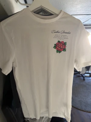 Vit t-shirt med ros och text - Vit t-shirt med tryck av en stor röd ros och texten 'Venice Beach Los Angeles' på ryggen. På bröstet finns en mindre ros och texten 'Endless Paradise'. T-shirten har rund hals och korta ärmar. Perfekt för dig som gillar streetwear och grafiska tryck.