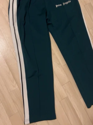 Palm Angels gröna mjukisbyxor - Mörkgröna mjukisbyxor från Palm Angels med vita och svarta sidoränder. Byxorna har resår i midjan och loggan broderad framtill. Tillverkade i ett mjukt och följsamt material som känns skönt mot huden. Perfekta för en avslappnad och trendig look. Pris kan diskuteras 