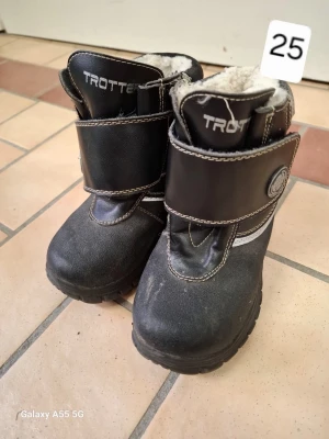 Svarta vinterstövlar från Trotter - Svarta vinterstövlar från Trotter med grov sula och bred kardborrestängning. Insidan är fodrad med mjukt vitt foder för extra värme. Stövlarna har vita kontrastsömmar och en rundad tå. Perfekta för kalla dagar.