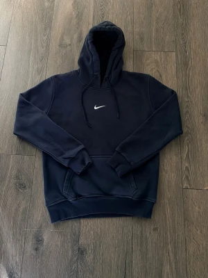 Svart vintage Nike hoodie med huva - Snygg svart vintage hoodie från Nike med klassisk vit swoosh-logga på bröstet. Hoodien har en stor huva med dragsko och en praktisk magficka. Tillverkad i mjukt och bekvämt material, perfekt för en avslappnad stil.