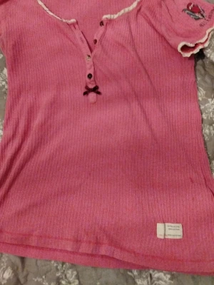 Rosa ribbad topp från Odd Molly - Supersöt rosa ribbad topp från Odd Molly med knappar framtill, liten rosett och dekorativ spetskant vid halsen. Kortärmad modell med broderad detalj på ärmen. Perfekt för dig som gillar en feminin och avslappnad stil.