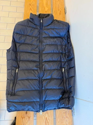 Mörkblå dunväst från Gant - Snygg mörkblå dunväst från Gant med quiltad design och hög krage. Västen har två dragkedjeförsedda fickor framtill och diskret logga på bröstet. Perfekt att slänga över en hoodie eller tröja för extra värme. Klassisk och stilren look.