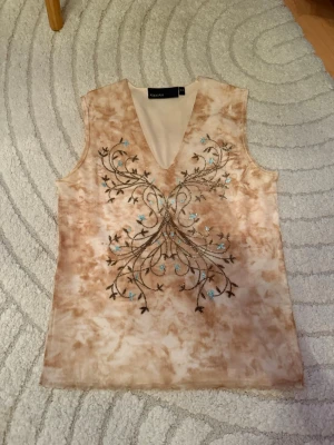 Beige ärmlös topp med broderi - Säljer en beige ärmlös topp från Rodday med v-ringning och broderade blommor i brunt och blått framtill. Toppen har ett unikt mönster och är perfekt för dig som gillar detaljer. Materialet känns mjukt och lätt, vilket gör den skön att bära.