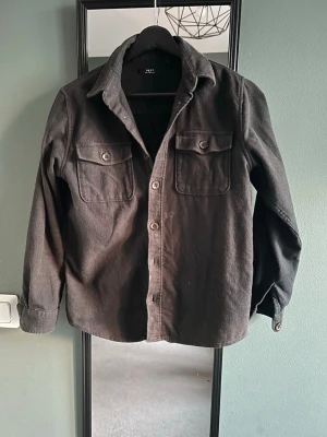  overshirt  - Stilren svart overshirt från NXT med två bröstfickor med lock och knapp, klassisk krage och knäppning framtill. Overshirten är tillverkad i ett mjukt tyg som ger en avslappnad look och passar perfekt till lager-på-lager-stil.