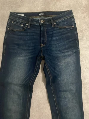 Jeans Jack & Jones - Snygga mörkblå slim fit jeans från Jack & Jones med klassisk femficksmodell och kontrastsömmar. Jeansen har lätt slitna detaljer framtill och är tillverkade i stretchigt denimtyg för extra komfort. Perfekta för en trendig och avslappnad stil.