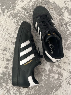 Adidas Superstar svartvita sneakers - Klassiska Adidas Superstar sneakers ( gold edition)  i svart läder. De är inte i perfekt skick eftersom har haft de i ca 8 månader, hälkappan är slitna annars ser de helt okej ut! Möts endast upp i Malmö, fraktar ej, priset går att förhandla vid snabbt köp! Säljer pga de är för små för mig! 