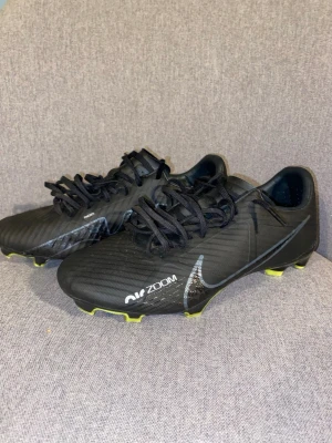 Nike Zoom svarta fotbollsskor - Säljer ett par svarta Nike Zoom fotbollsskor med snörning och låg profil. Skorna har en stilren design med subtila detaljer och Nike-logga på sidan. Sulan har gula dobbar för bra grepp på planen. Perfekta för dig som vill ha både stil och funktion på fotbollsplanen.