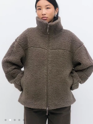 Brun teddyjacka med hög krage - Mysig brun teddyjacka med hög krage och hel dragkedja framtill. Jackan har en oversized passform och är gjord i fluffigt teddy-material som ger en trendig look. Perfekt för att hålla värmen under kyliga dagar. Inga synliga loggor eller märken.