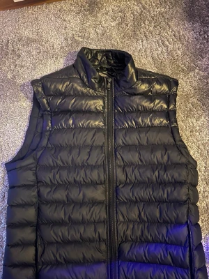 Svart dunväst från J.Lindeberg - Snygg svart dunväst från J.Lindeberg med hög krage och dragkedja framtill. Västen har en quiltad design med horisontella sömmar och är tillverkad i glansigt material. Perfekt för lager på lager och ger en clean look. Köpt i j lindebergs butik i Stockholm. Nypris 1800, säljer då den ej kommer till användning. Skriv om du har några frågor eller funderingar!