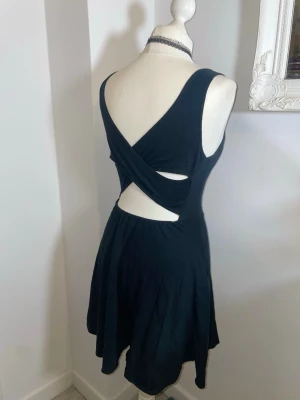 Svart kort klänning med öppen rygg - Svart kort klänning med rund halsringning och snyggt korsad öppen rygg. Klänningen är ärmlös och har en figurnära passform upptill med utställd kjol. Perfekt för dig som gillar stilrena och trendiga plagg.