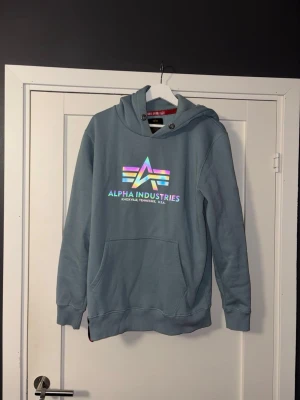Blå hoodie Alpha Industries L - Snygg blå hoodie från Alpha Industries med stor ficka framtill och huva med metallöljetter. Framtill finns en färgglad logga och texten 'Alpha Industries'. Mjuk insida och ribbade muddar vid ärmar och nederkant. Perfekt för en avslappnad streetwear-look.