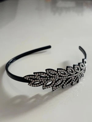 Svart diadem med glittriga stenar - Snyggt svart diadem dekorerat med gnistrande strasstenar i ett bladliknande mönster. Perfekt accessoar för att lyfta din look med lite extra bling. Diademet är smalt och flexibelt, vilket gör det bekvämt att bära hela dagen.
