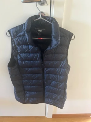 Mörkblå dunväst från Uniqlo - Stilren mörkblå dunväst från Uniqlo med lättviktigt material och quiltad design. Västen har hög krage och dragkedja framtill, perfekt för lager-på-lager och enkel att matcha med olika outfits. 
