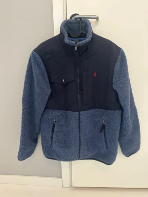 Blå teddyjacka från Polo Ralph Lauren, nyskick - Mysig blå teddyjacka från Polo Ralph Lauren med kontrasterande mörkblå partier på axlar och bröst. Jackan har hög krage, dragkedja framtill, två sidofickor med dragkedja och en bröstficka med knapp. Röd logga broderad på bröstet. Perfekt för kalla dagar.