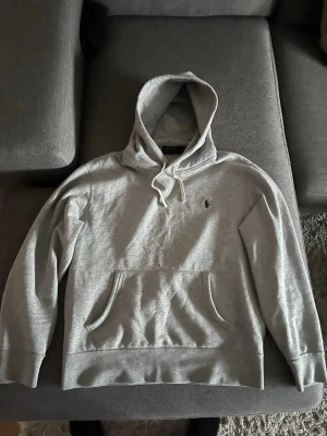 Grå hoodie från Polo Ralph Lauren - Snygg grå hoodie från Polo Ralph Lauren med klassisk huva och dragsko. Har en känguruficka framtill och den ikoniska lilla loggan broderad på bröstet. Tillverkad i mjukt bomullsmaterial som är perfekt för chill dagar.