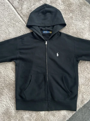 Svart hoodie från Polo Ralph Lauren - Snygg svart hoodie med dragkedja från Polo Ralph Lauren. Klassisk design med vit broderad logga på bröstet, två fickor fram och skön huva. Tillverkad i mjukt bomullsmaterial som känns riktigt bekvämt. Perfekt för en avslappnad streetwear-look.