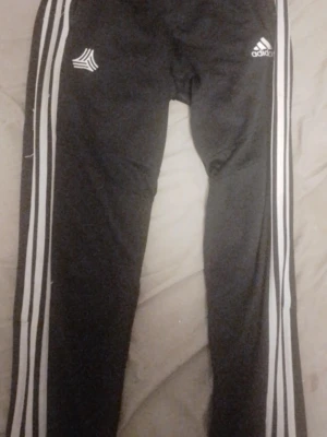 Svarta Adidas träningsbyxor - Säljer ett par svarta träningsbyxor från Adidas med de klassiska vita ränderna längs benen och Adidas-logga på låret. Byxorna har smal passform och är gjorda i ett stretchigt material som passar perfekt för sport och träning. Lite slitna vid dom vitna ränderna (kolla bild