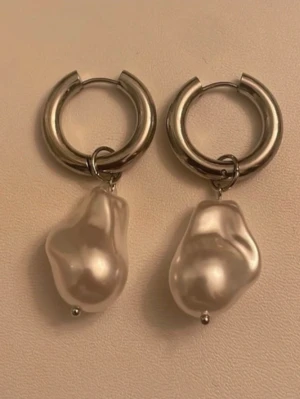 Silvriga hoops med oregelbundna pärlor - Snygga örhängen med tjocka silvriga hoops och stora, oregelbundna vita pärlor som hänger ned. Perfekt för dig som gillar statement-smycken med en modern twist. Materialet ser ut att vara metall och konstpärlor.
