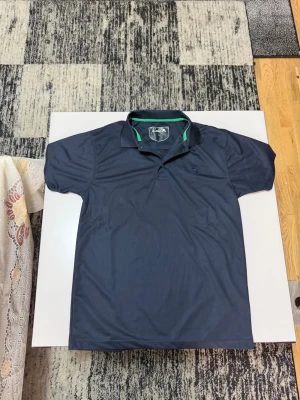 Mörkblå piké från Arnold Palmer - Snygg mörkblå pikétröja från Arnold Palmer med klassisk krage och grönt kantband på insidan. Tröjan har korta ärmar och en diskret logga på bröstet. Perfekt för en avslappnad men stilren look.