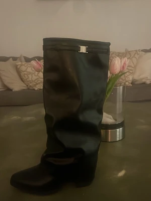 Svarta slouchy boots med metallspänne helt nya - Säljer ett par svarta boots i slouchy modell med rund tå och ett stilrent metallspänne upptill. Skorna har en mjuk, glansig yta som ger en cool look och passar perfekt till jeans eller kjol. Materialet ser ut att vara konstläder.anledning varför ja säljer för liten storlek