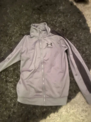 Grå ziptröja från Under Armour - Snygg grå ziptröja från Under Armour med hög krage och hel dragkedja. Tröjan har mörkare grå detaljer längs ärmarna och logga på bröstet. Perfekt för träning eller chill. Tillverkad i mjukt och stretchigt material.