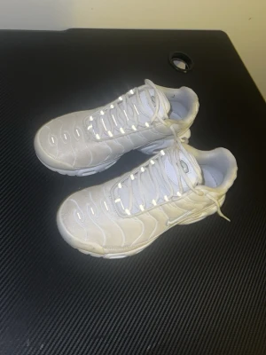 Nike Air Max Plus vita sneakers - Nike Air Max Plus sneakers i helvitt med mesh och syntetpaneler. Skorna har den ikoniska vågiga designen, synlig Air-enhet i sulan och vita skosnören. Swoosh-logga på sidan och TN-logga på hälen. Perfekta för dig som gillar sportig och clean stil.