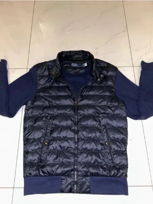Mörkblå cardigan jacka från Polo Ralph Lauren - Snygg mörkblå cardiganjacka från Polo Ralph Lauren med quiltad, glansig front och mjuka stickade ärmar och rygg. Jackan har dragkedja, ståkrage och två fickor med knappstängning. Perfekt för lager på lager och en sportig, modern look.