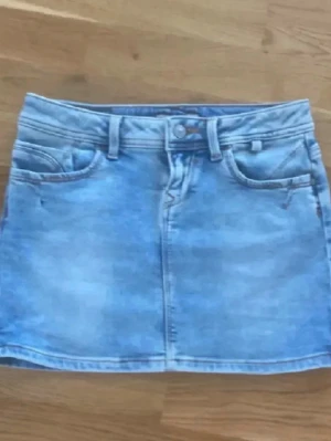 Ljusblå jeanskjol med fickor - Säljer en ljusblå jeanskjol i klassisk modell med fem fickor, bälteshällor och knappstängning fram. Kjolen har en kort passform och är tillverkad i denim med snygga sömdetaljer både fram och bak.