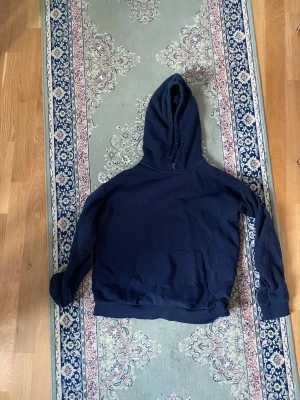 Mörkblå hoodie med huva - En enkel och stilren mörkblå hoodie med stor ficka framtill och justerbar huva. Hoodien har en avslappnad passform och ribbade muddar vid ärmar och nederkant. Perfekt för en casual look och lätt att matcha med andra plagg.