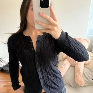 Svart kabelstickad kofta - Svart kabelstickad kofta med rund halsringning och knäppning framtill. Långa ärmar och ribbade muddar vid ärmslut och nederkant. Perfekt för lager-på-lager och enkel att matcha med jeans eller kjol.