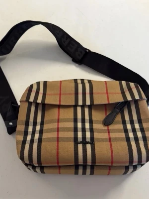 Burberry axelväska i beige rutigt tyg - Snygg axelväska från Burberry i klassiskt beige rutigt mönster med svarta, vita och röda ränder. Väskan har justerbar svart axelrem med logga, lock med spänne och dragkedja. Perfekt för dig som vill ha en trendig och ikonisk accessoar.