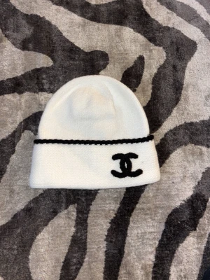 Vit mössa från Chanel med logga - Säljer en stilren vit mössa från Chanel med svart broderad logga framtill och en svart detalj längs kanten. Mössan är mjuk och stickad, perfekt för att hålla värmen och samtidigt se trendig ut. Klassisk design som passar till många olika outfits.